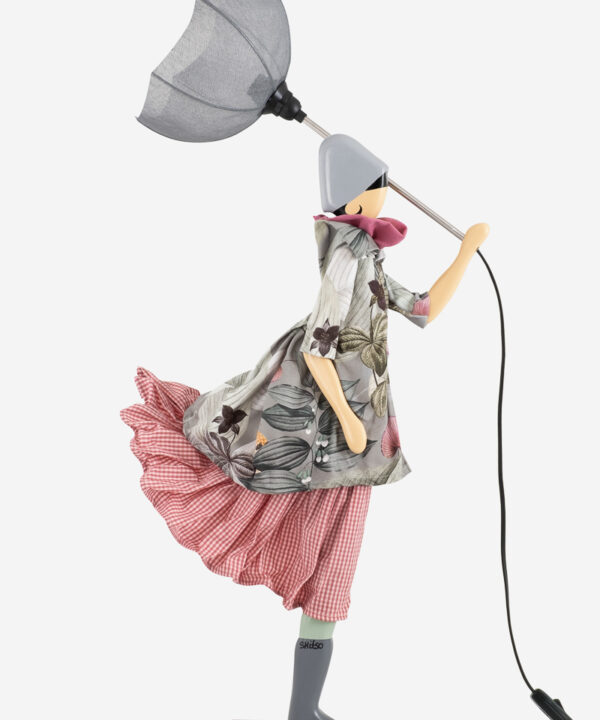 En tilsvarende lampe med en figur, der holder en grå paraply, iført en kjole med en blanding af blomster- og ternmønster. Kjolen kombinerer afdæmpede og klare farver, hvilket skaber en balance mellem elegance og legesyge.